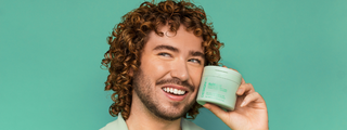 A smiling man with coily brown curls holding the GLOSS.ME hydrating treatment mask against a green background.