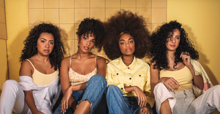 Four women with natural curly and coily hair, sitting side by side against a warm yellow tiled wall, showcasing different curl patterns and volume