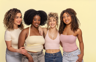 Four women with different hairstyles posing together and smiling against a yellow background.