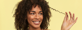 Woman with coily hair holding a section of it against a yellow background.