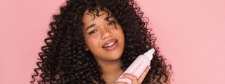 A woman with coily hair smiling and holding the PUFF.ME Mousse styling product against a pink background.