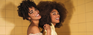 Two women with voluminous natural curly and coily hair smiling against a yellow tiled background.