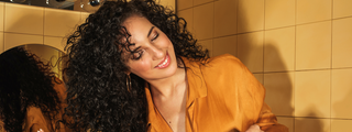 A woman with coily hair, smiling and wearing an orange shirt against an orange tiled background.