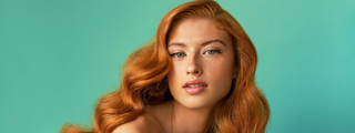 Portrait of a woman with long, wavy, vibrant auburn waves and blue eyes against a mint green backdrop.