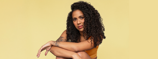 A woman with voluminous, coily dark curls and a floral arm tattoo posing against a soft yellow background.