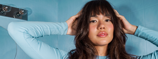 A woman with long dark hair and a soft fringe posing with her hands to her head in a blue-tiled room.