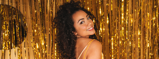 Smiling woman with voluminous dark curls posing in front of a shiny gold tinsel backdrop.