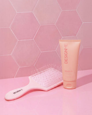 DESIGNME detangling paddle brush and HOLD.ME styling cream sit side-by-side with a pink tiled background.